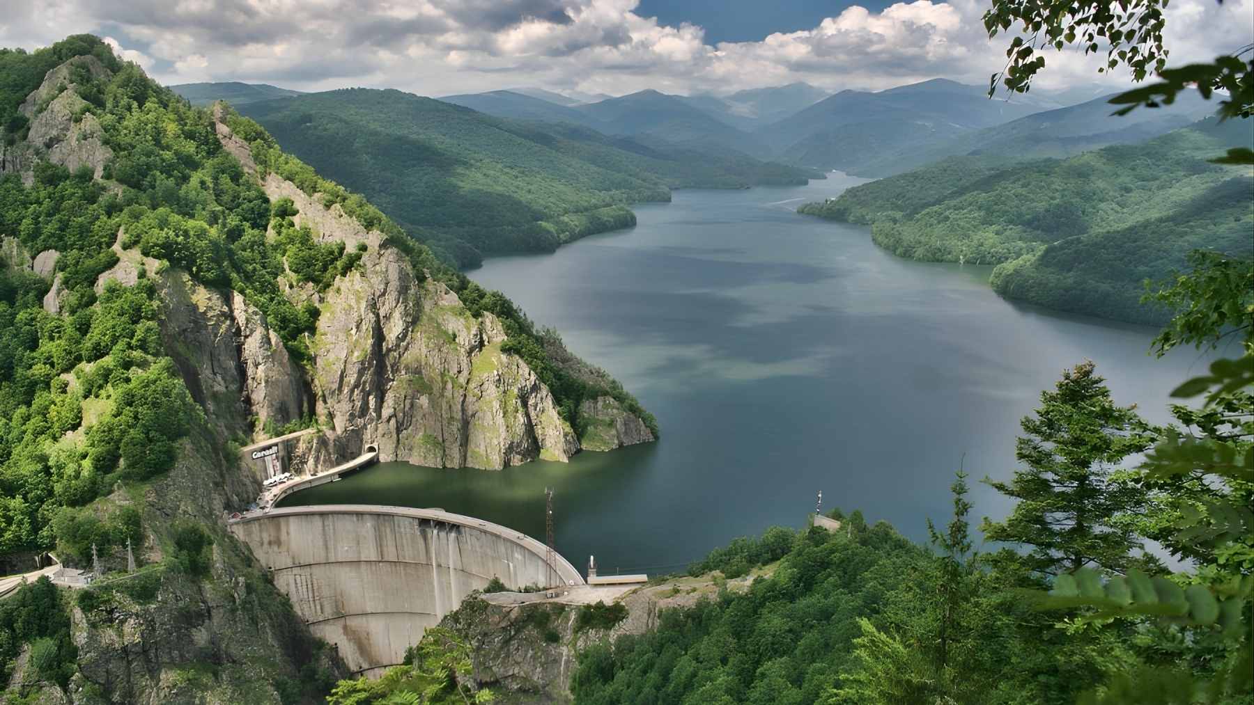 Mountain reservoir and hydroelectric dam in Romania’s Carpathians, with a curved concrete wall holding back a long lake between green ridges.