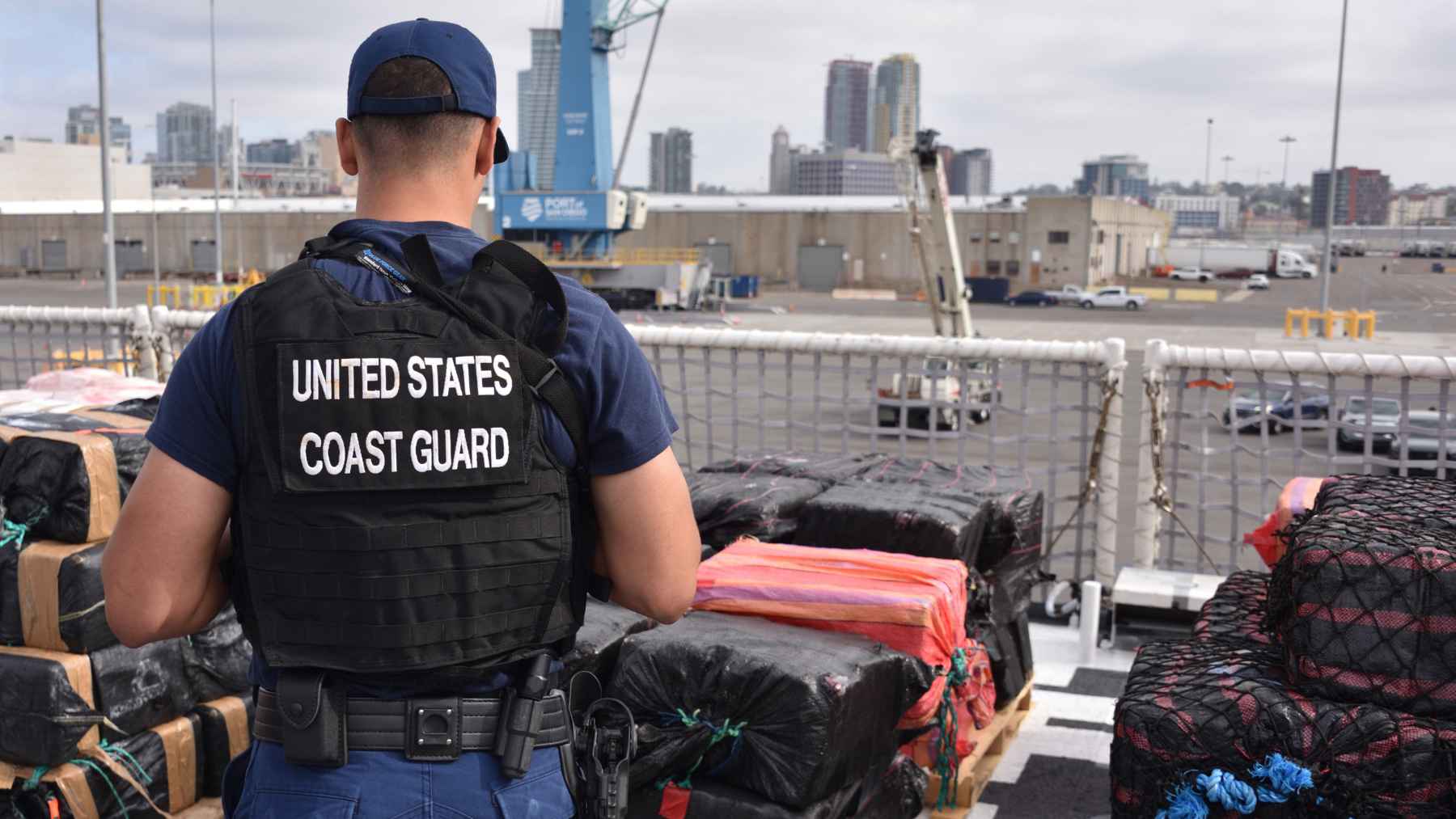A U.S. Coast Guard member stands beside stacks of seized cocaine bales during an offload at a port.
