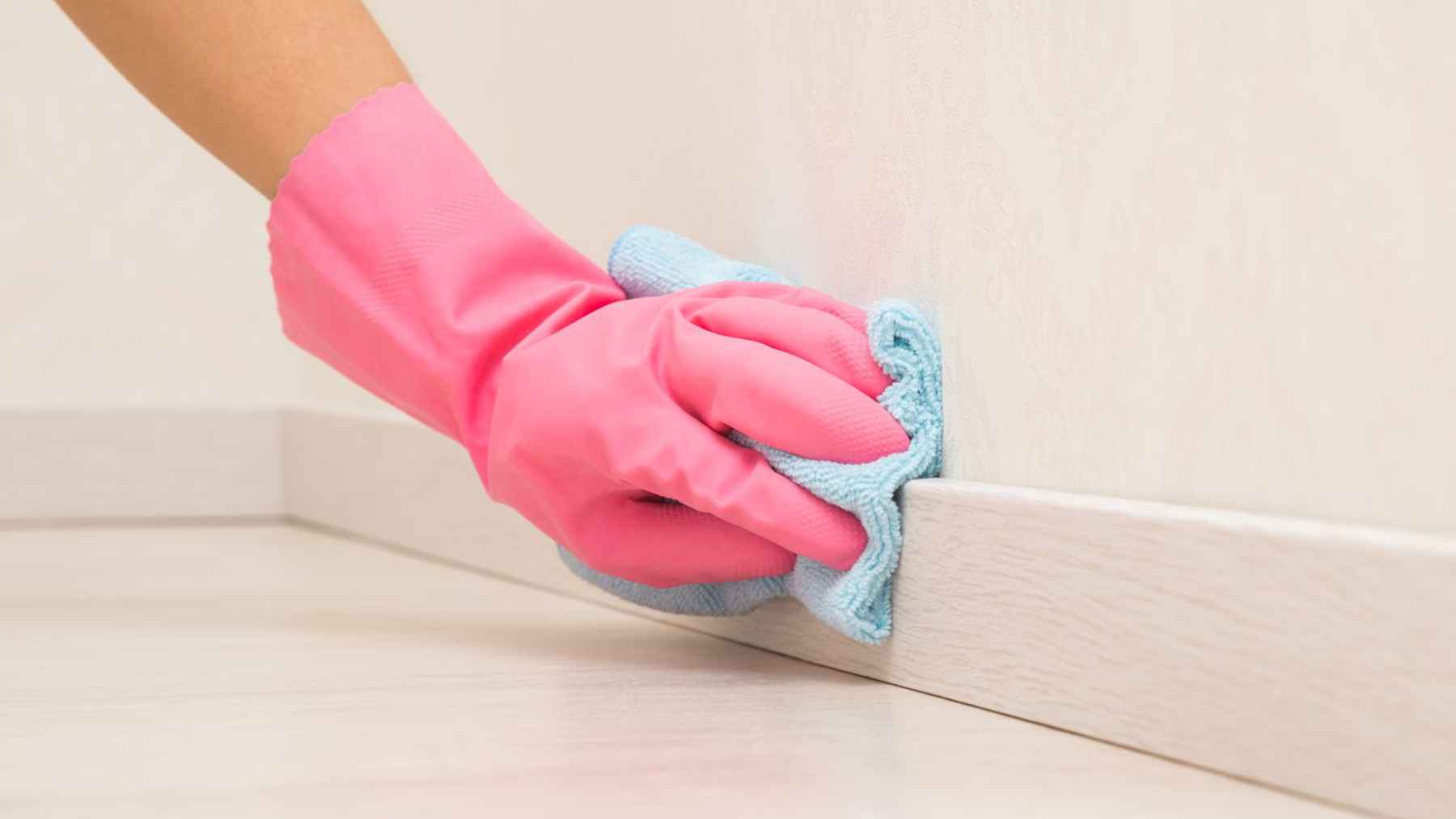 A hand in pink cleaning gloves wipes a baseboard with a microfiber cloth, showing a simple trick to keep dust from building up.