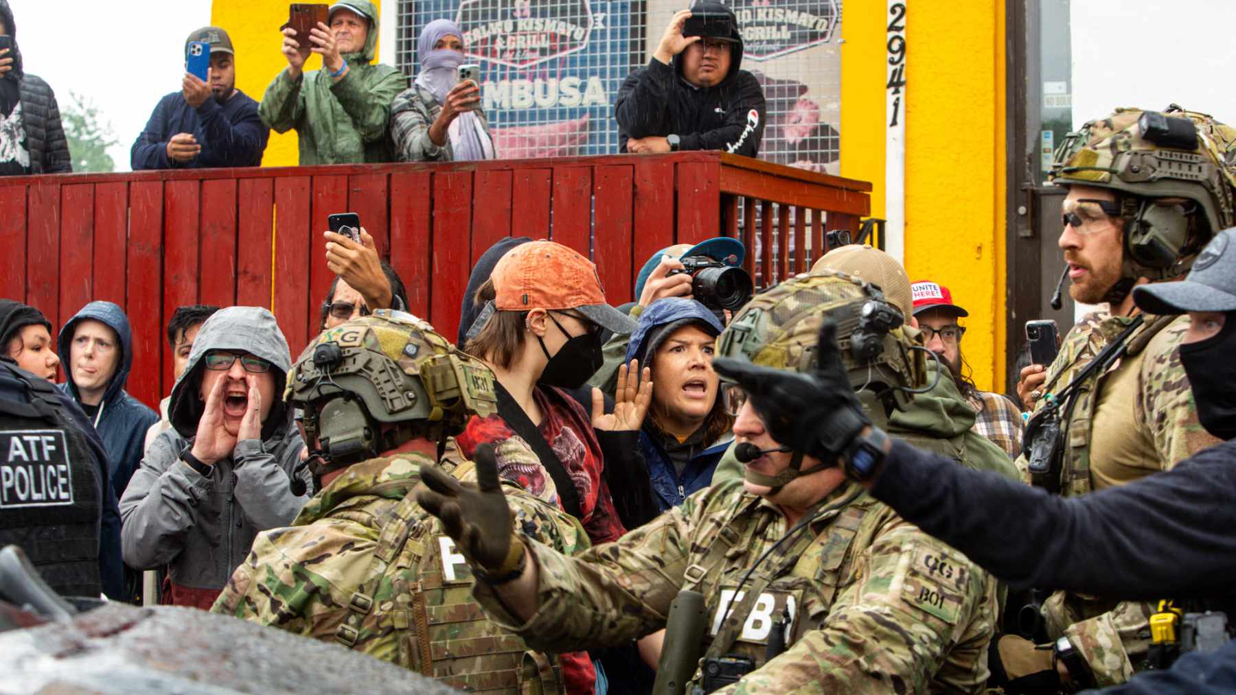Federal agents in tactical gear face a crowd filming with phones during a tense operation outside a building in Minneapolis.
