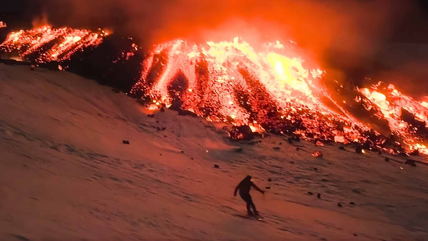 A skier descends a snowy slope on Mount Etna as a lava glow and thick ash plume rise behind the ridge in eastern Sicily.