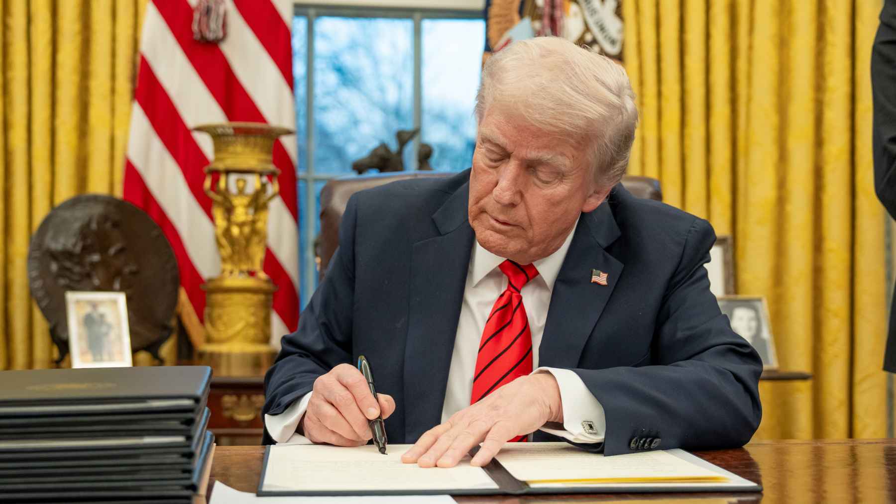A US president signs an emergency order at the Resolute Desk as aides stand nearby in the Oval Office.