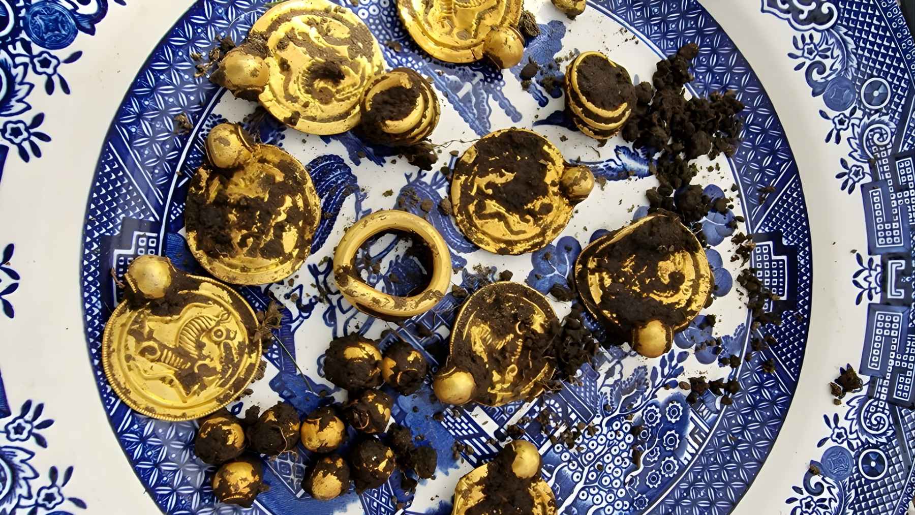 Gold bracteate pendants, beads, and rings covered in soil on a patterned plate after being recovered from a Norwegian archaeological find.