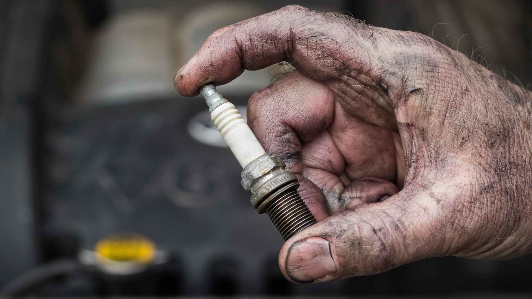 Grease-stained hand holding a spark plug in front of a car engine bay.