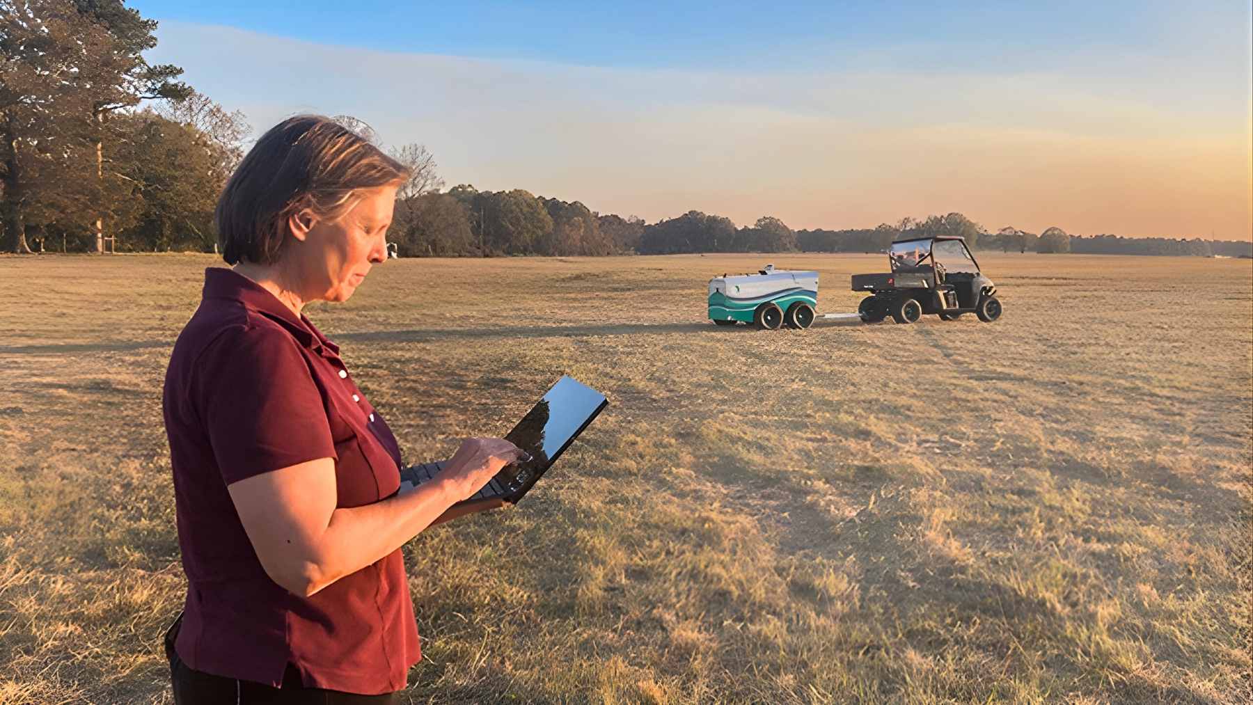 Researcher uses a laptop in a grassy field while a utility vehicle and equipment sit in the background