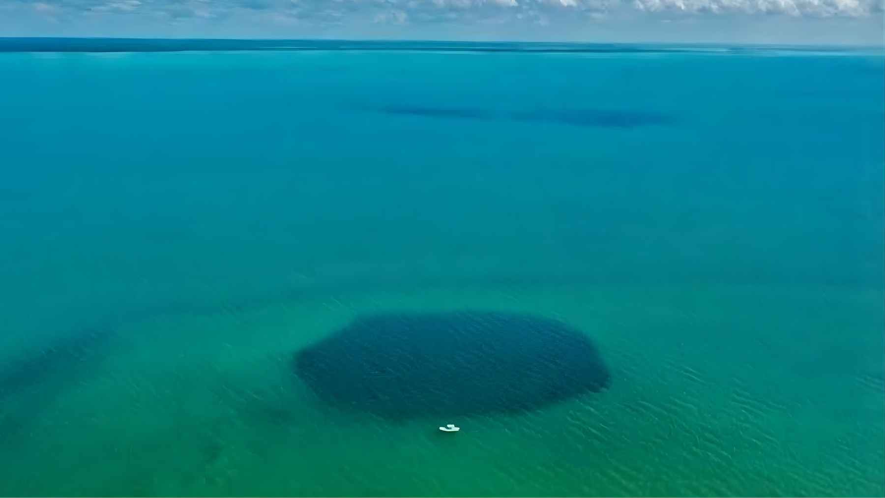 A dark circular patch marks the Taam Ja blue hole in Chetumal Bay, Mexico, seen from above with a small boat nearby.