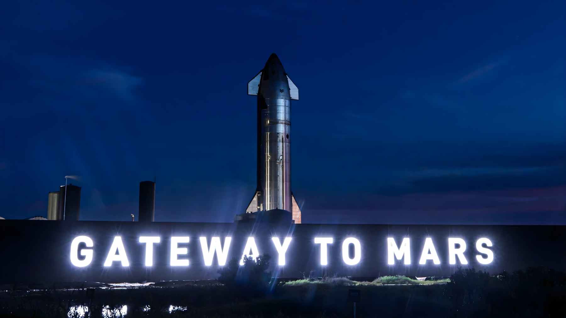SpaceX Starship stands behind a glowing “Gateway to Mars” sign at Starbase in South Texas at night.