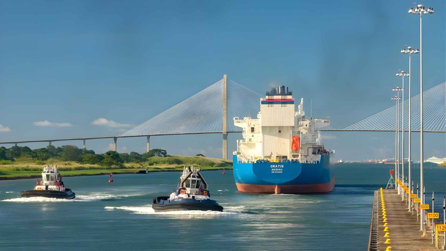 A cargo ship escorted by tugboats moves through a canal channel with a bridge in the background.