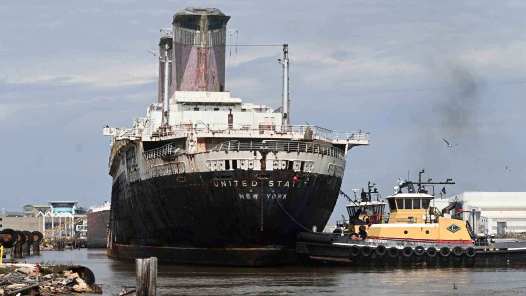 The SS United States ocean liner sits in port as tugboats guide it during a tow, beginning its journey to become an artificial reef.