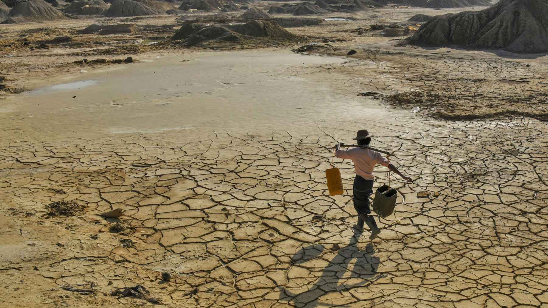 Person walking across cracked dry land carrying water containers, illustrating worsening drought and future global water shortages.