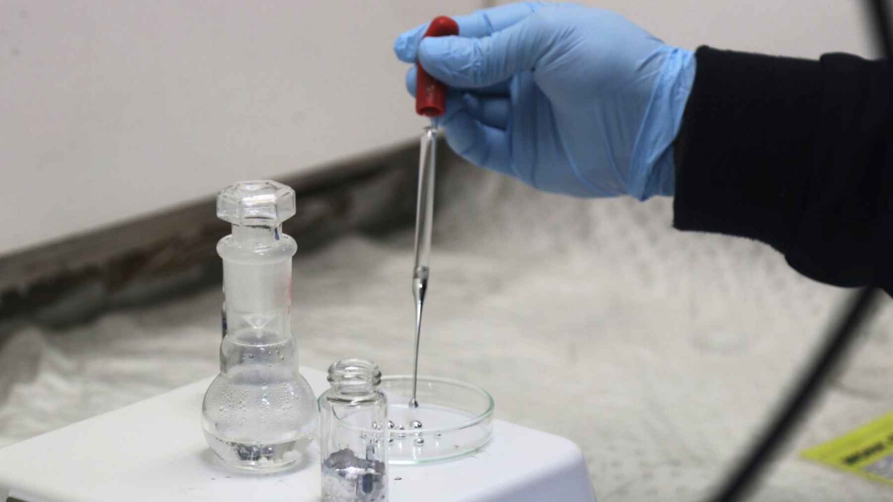 Gloved hand using a pipette in a lab setup with gallium and glassware, illustrating hydrogen production from sunlight and water