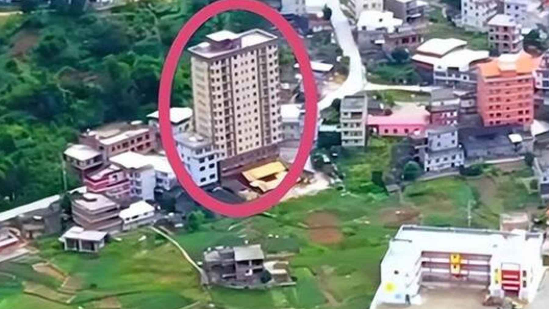 Aerial view of a 15-story tan apartment tower standing prominently in the center of a green, rural Chinese village.