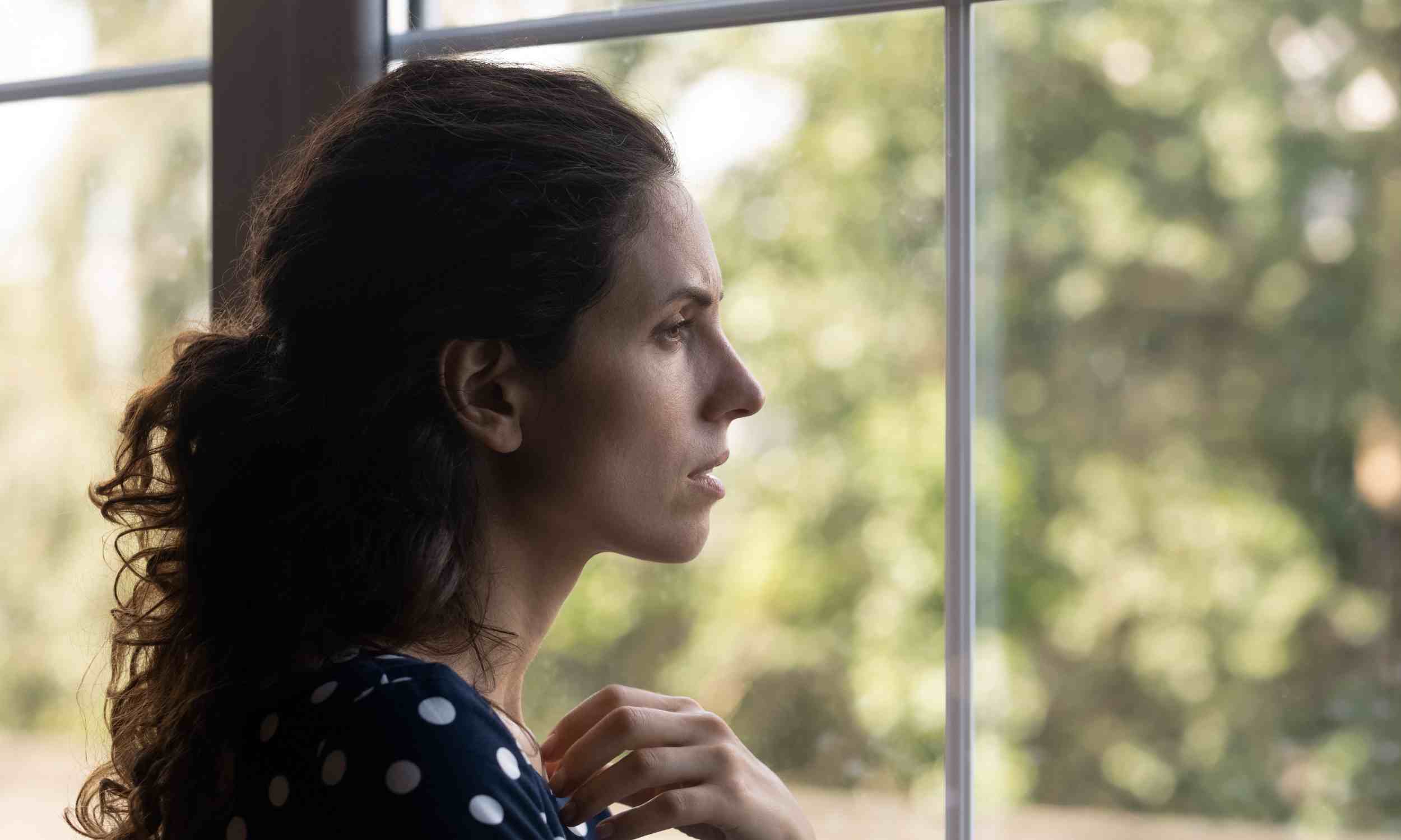 Woman looking out a window alone, illustrating why kind people may struggle to form close friendships, according to psychology
