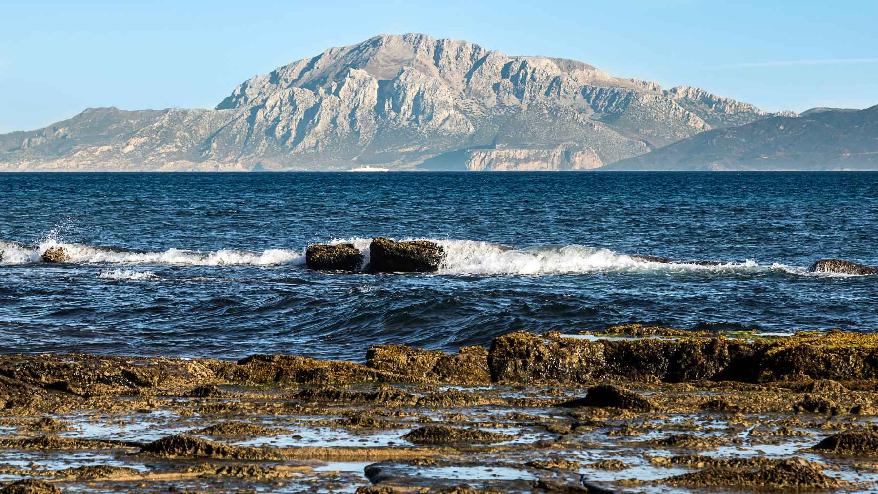 Rocky shoreline and sea view facing a large mountain across the Strait of Gibraltar, illustrating the tectonic zone linked to Iberia’s slow geological movement.