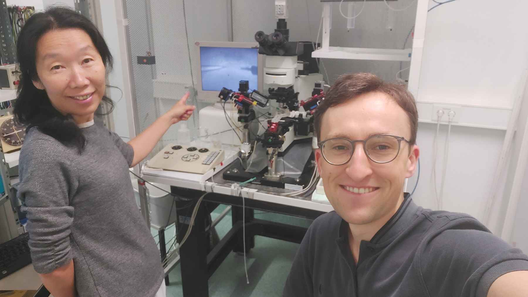 Two researchers smiling in a laboratory setting, with one pointing to a microscope and computer monitor displaying cryopreserved brain cells.