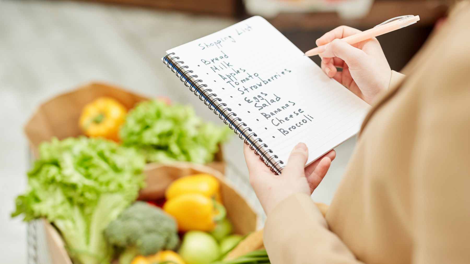 A handwritten grocery list on a piece of lined paper next to a pen and a shopping basket, emphasizing the physical act of planning.
