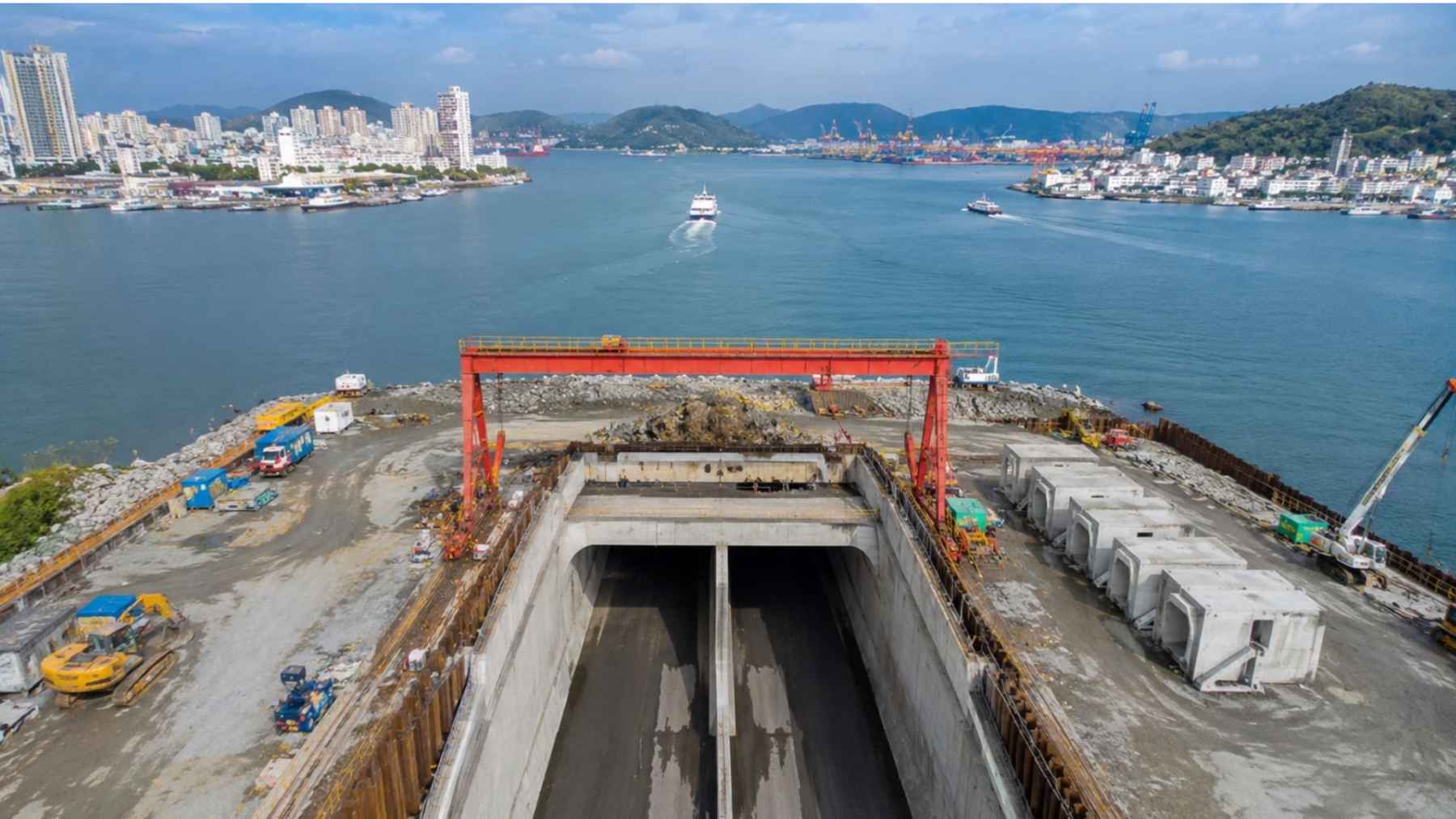 Construction site for an immersed tunnel beside a wide port channel, with concrete tunnel sections, cranes, boats, and city buildings in the background.