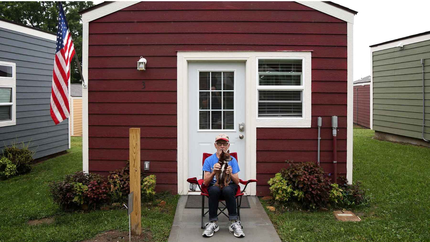 Plans are underway in Cincinnati to build a new “Veterans Village” featuring 14 tiny homes on church-owned land. Each unit, measuring 276 square feet, will include a porch, kitchen, dining area, and full bathroom, and will cost $70,000, allowing veterans to move out of their temporary housing 1 Veteran standing outside a tiny home with a dog on the porch, representing stable housing and daily life in a small community.