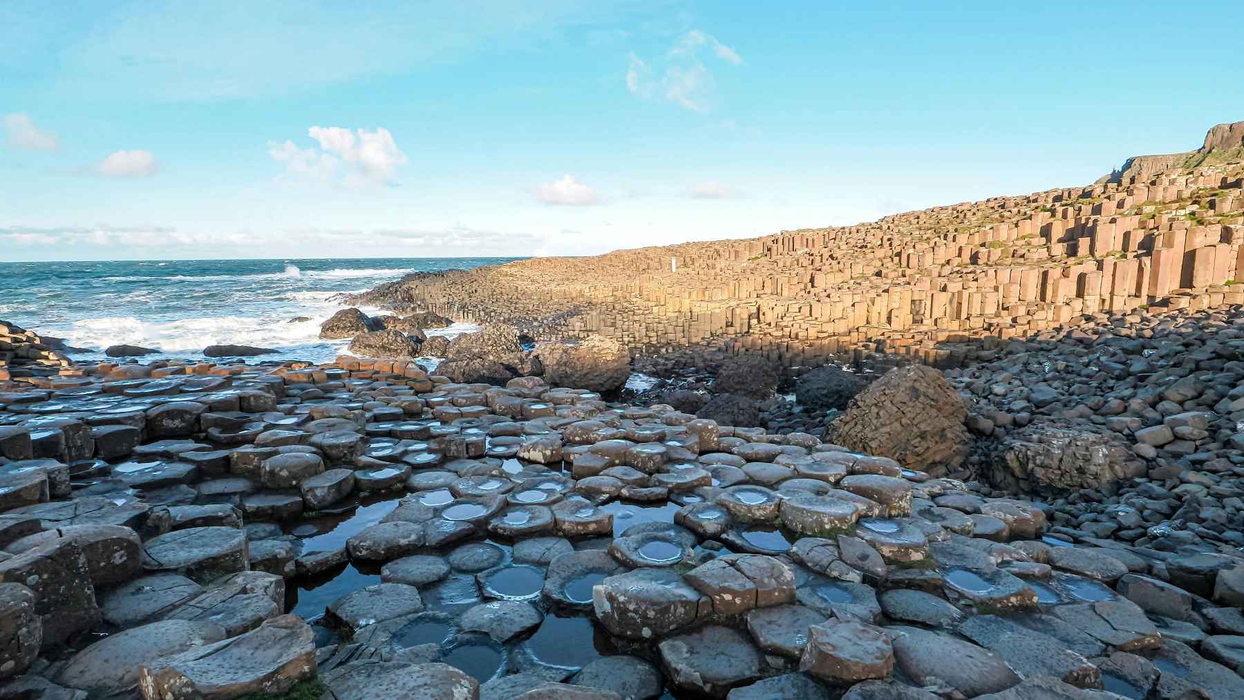 Wide coastal view of Giant’s Causeway with hexagonal basalt stones, tide pools, and Atlantic waves in Northern Ireland.