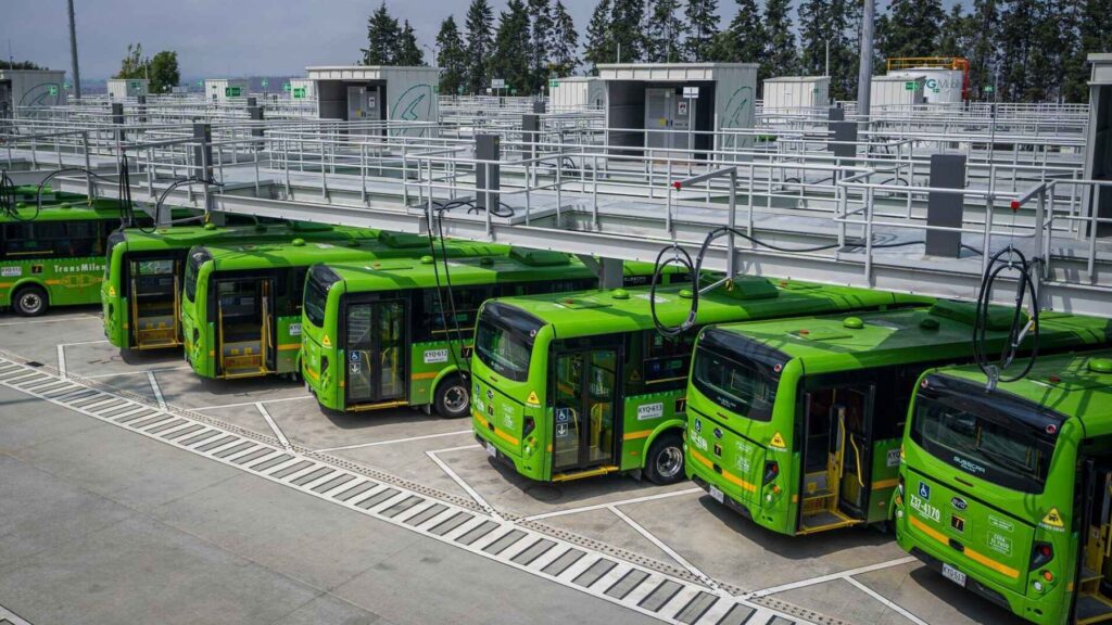 Row of green TransMilenio electric buses plugged into overhead chargers at Bogotá’s Calle Sexta depot.