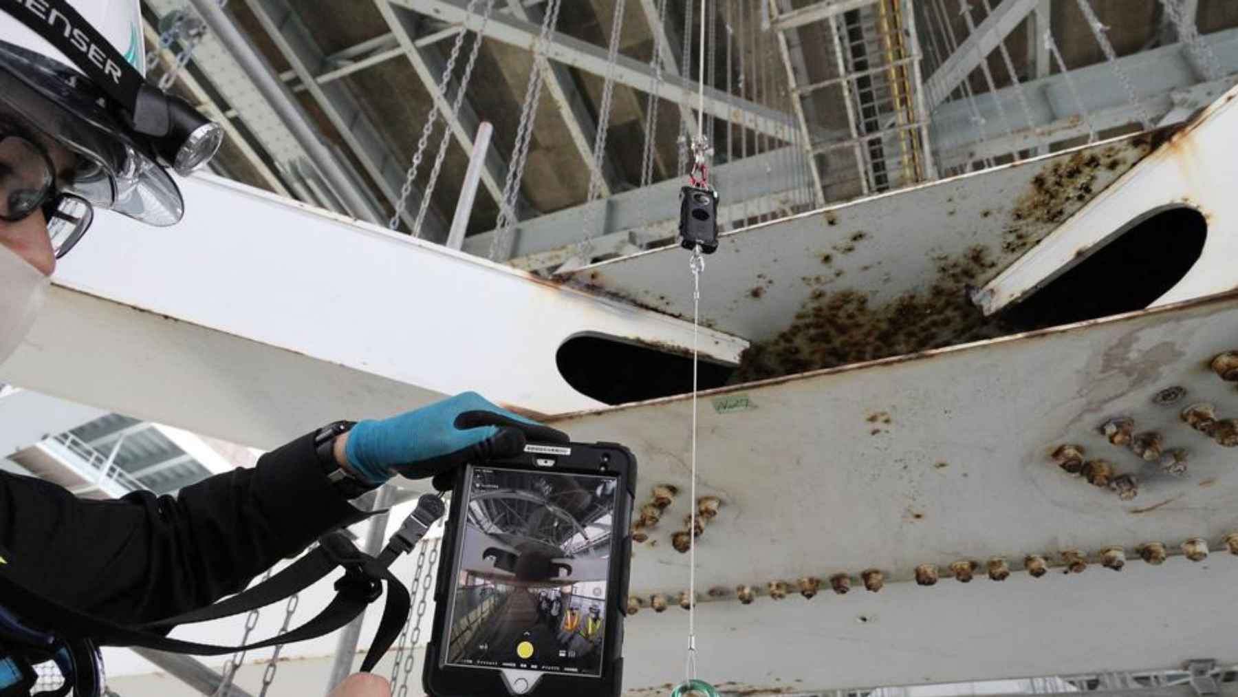 Close-up of corroded bridge steel with an inspector using a digital device to assess structural damage.