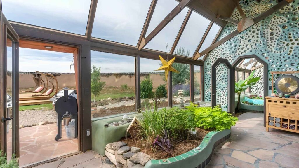 Indoor greenhouse inside an Earthship home with large solar-facing windows, food-growing beds, and recycled glass bottle walls.