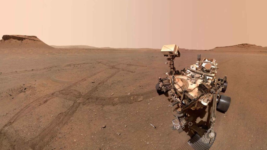 Wide-angle mosaic of Perseverance amid sweeping wheel tracks that curve across the floor of Jezero Crater.
