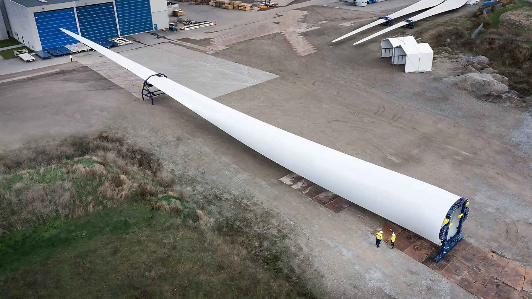 Full-size wind turbine blade lying at an industrial facility with two workers standing beside it.