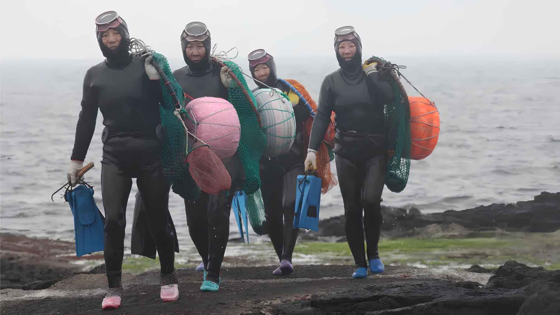 Jeju haenyeo women carrying diving gear, nets and colorful tewak floats beside the sea.