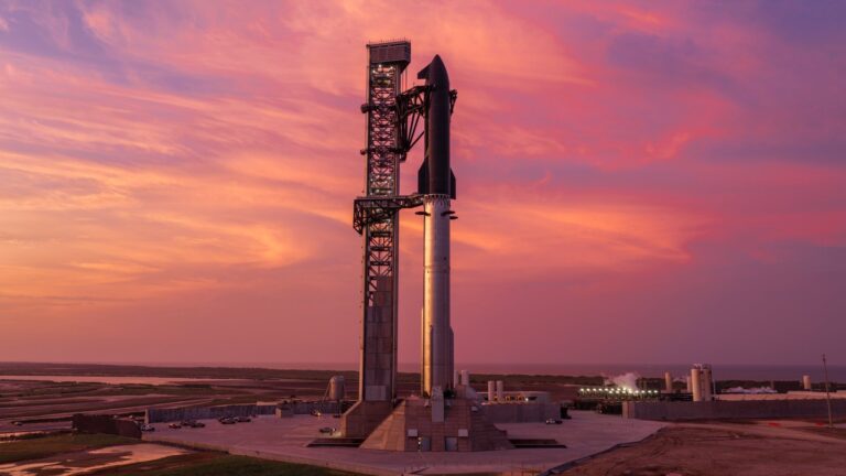 The full SpaceX Starship and Super Heavy stack standing beside the launch tower at Starbase under a pink and orange sunset sky