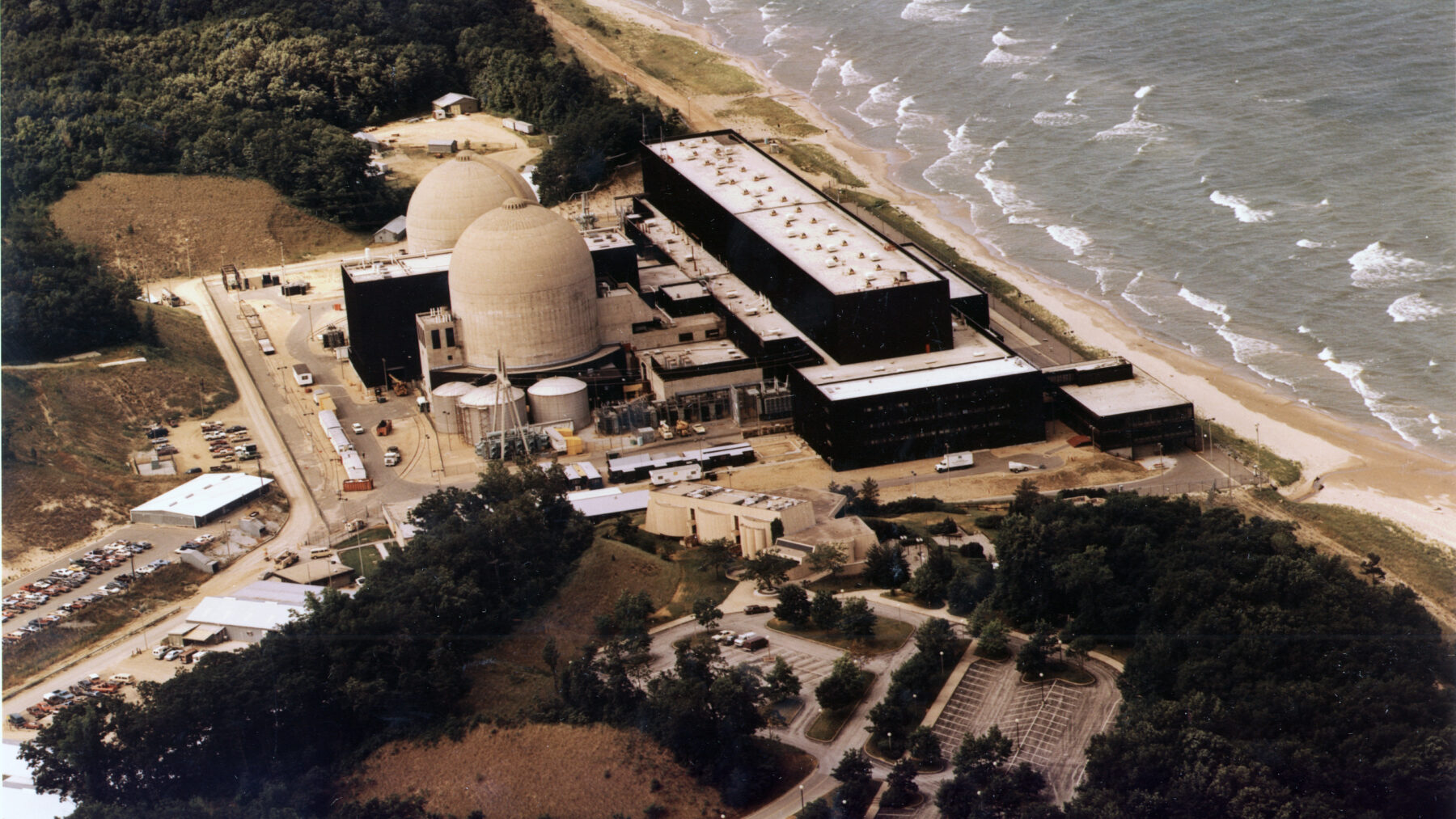 Aerial view of a nuclear power plant with two domed reactor buildings and long dark turbine halls beside the waves of Lake Michigan.