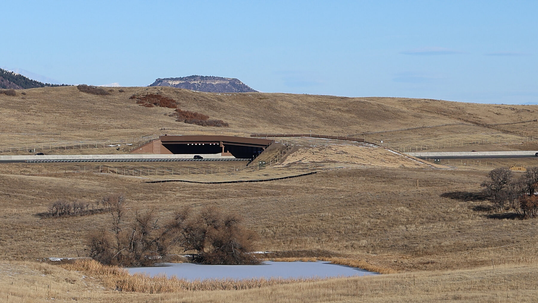 The Greenland Wildlife Overpass spanning Interstate 25 in Douglas County, Colorado, seen from across open grassland with a small pond in the foreground