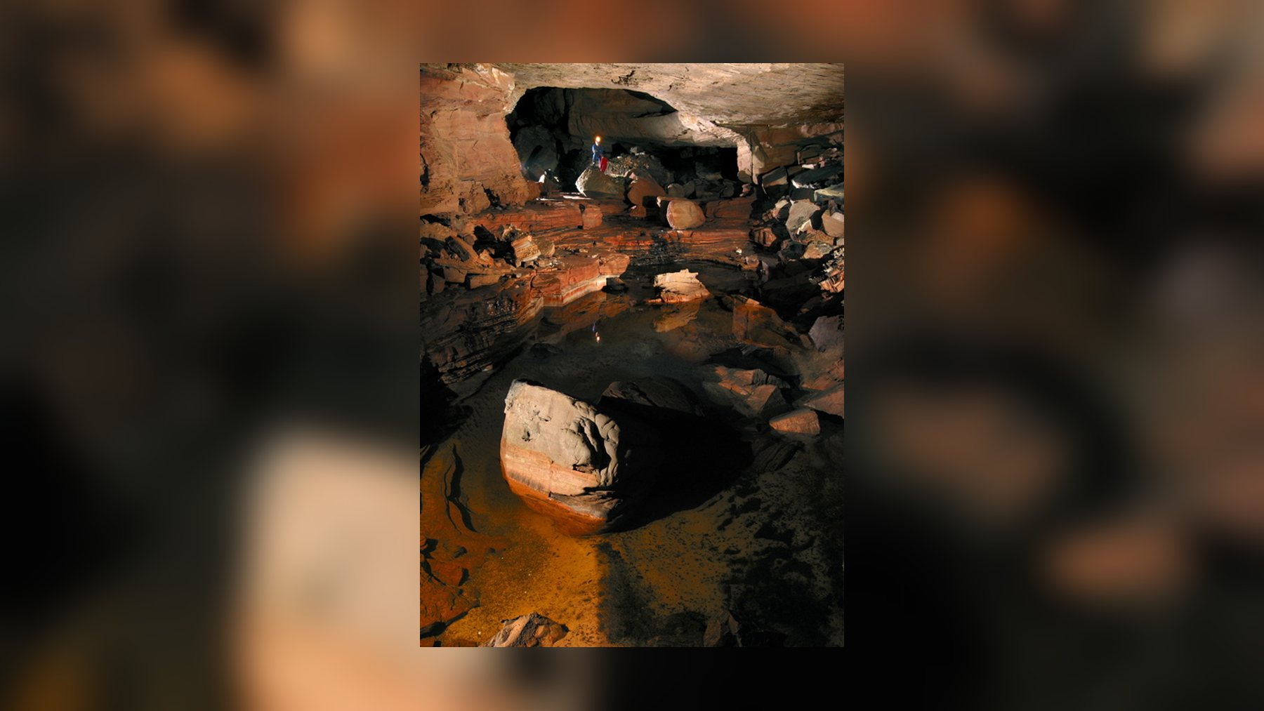 A caver with a headlamp stands at the back of a chamber with a pool and pink, layered rock in Cueva Ojos de Cristal, a quartzite cave on Mount Roraima