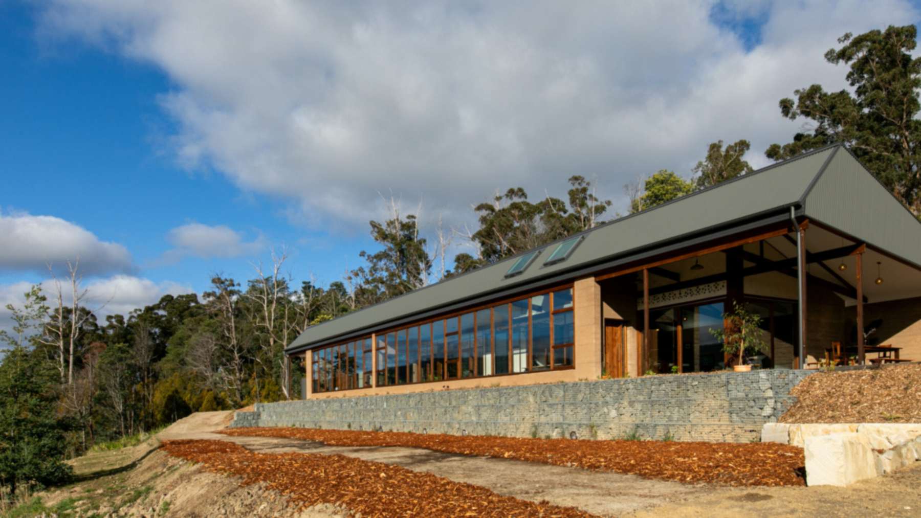 Cygnet Earthship in Tasmania with rammed earth walls, a long north-facing glass sunroom and a rock gabion wall below