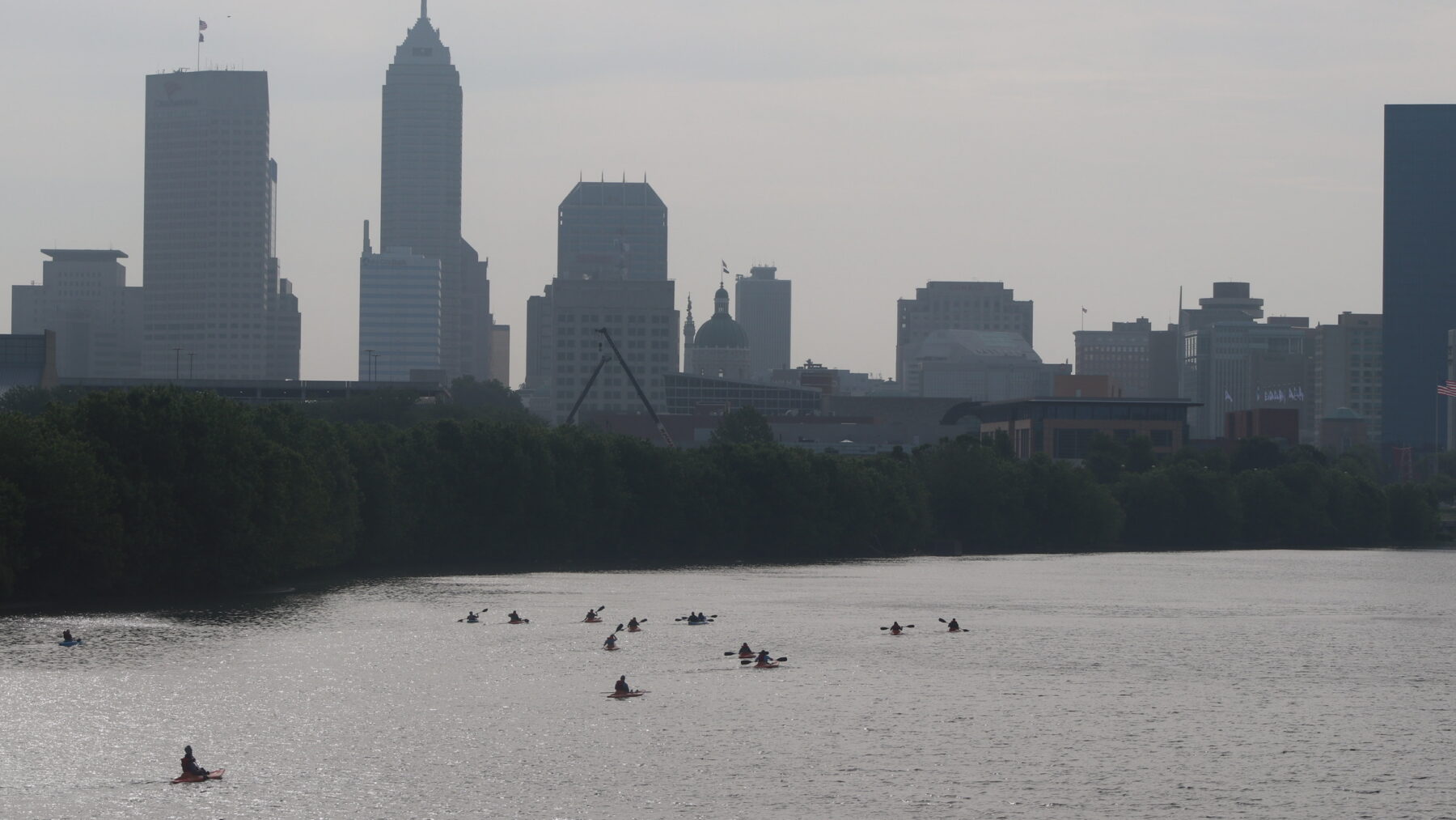 Kayakers paddle across the wide White River with the Indianapolis skyline rising through the haze behind them.