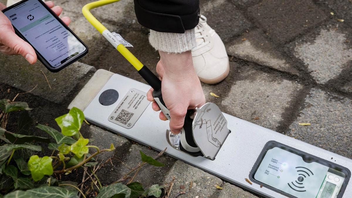 Hand pushing open the socket flap of a Rheinmetall curb charger with a charging plug while holding a phone