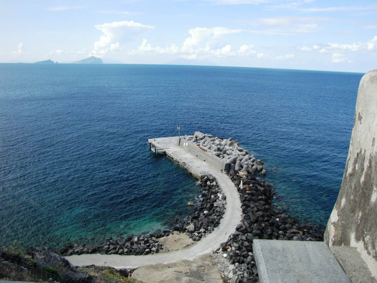 A small stone jetty in a cove below a cliff, with islands on the horizon