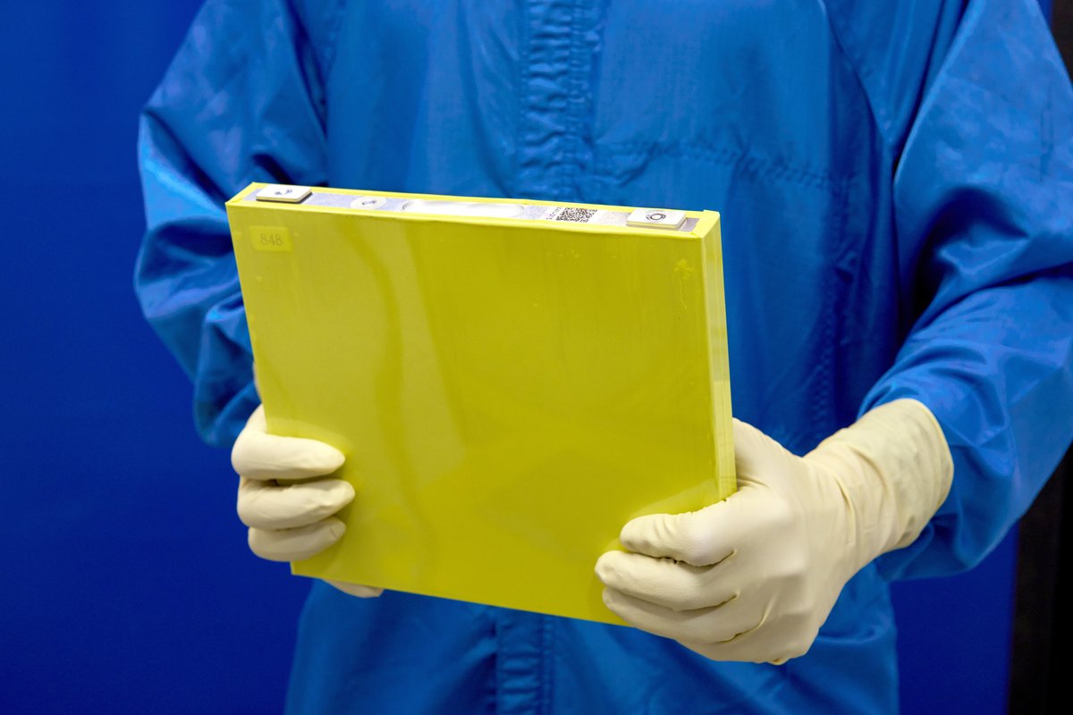 A gloved employee in a blue lab coat holds a large flat yellow battery cell