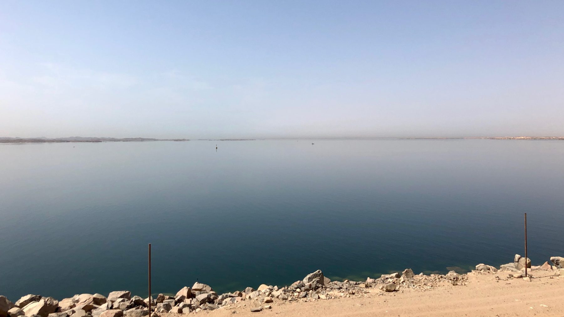 Calm blue water of Lake Nasser seen from the top of the Aswan High Dam, with a rocky shore and sandy road in the foreground