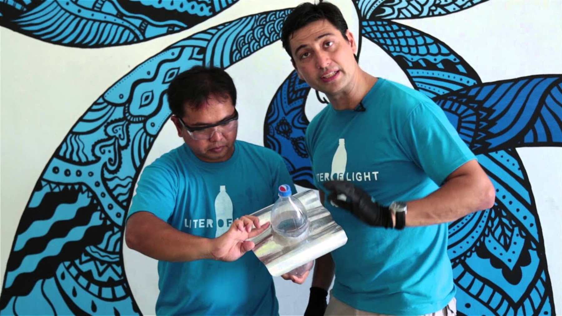 Two Liter of Light volunteers hold a water-filled solar bottle bulb fitted into a sheet of metal roofing