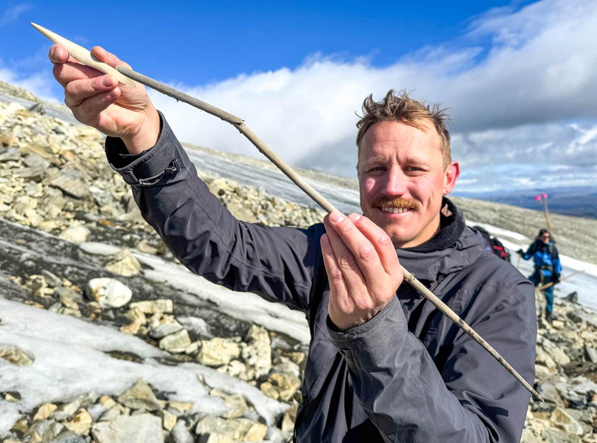 Archaeologist Kjetil Loftsgarden holding a complete prehistoric arrow with a bone tip and wooden shaft near an ice patch in Norway