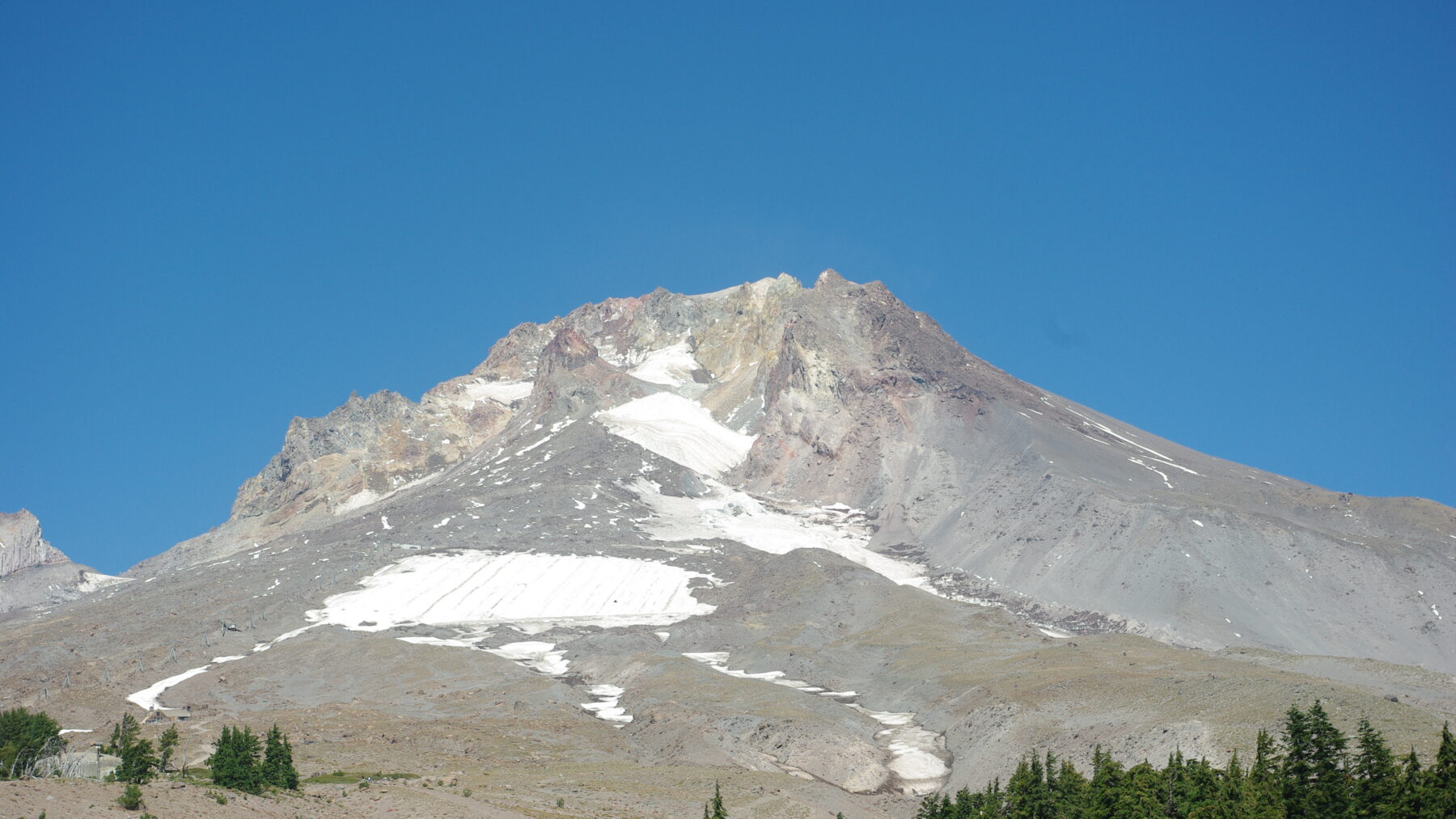 Bare, rocky upper slopes on the south side of Mount Hood in late summer, with a small patch of the Palmer Snowfield and a few conifers under a clear blue sky
