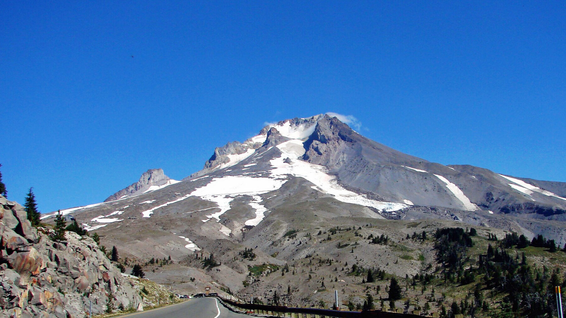 Mount Hood from a mountain road in September, its slopes mostly bare rock and gravel with only scattered patches of snow and ice under a deep blue sky