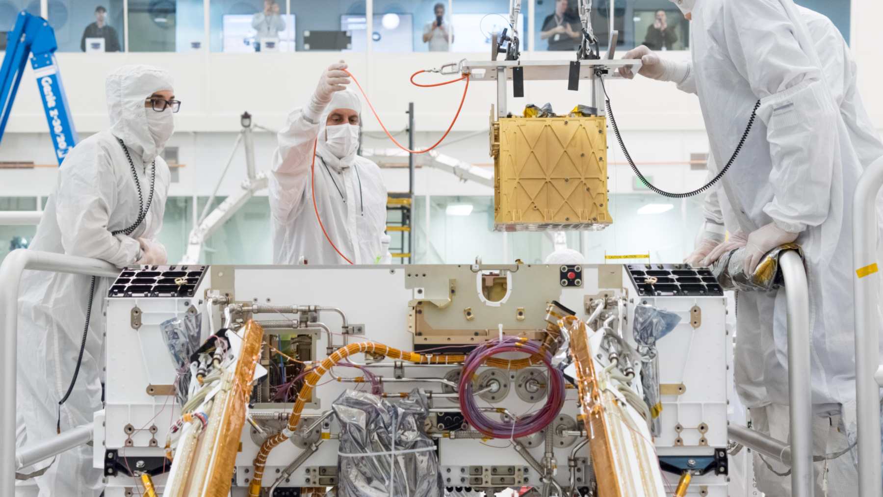 Engineers in a clean room lower the gold-colored MOXIE instrument into the Perseverance rover chassis