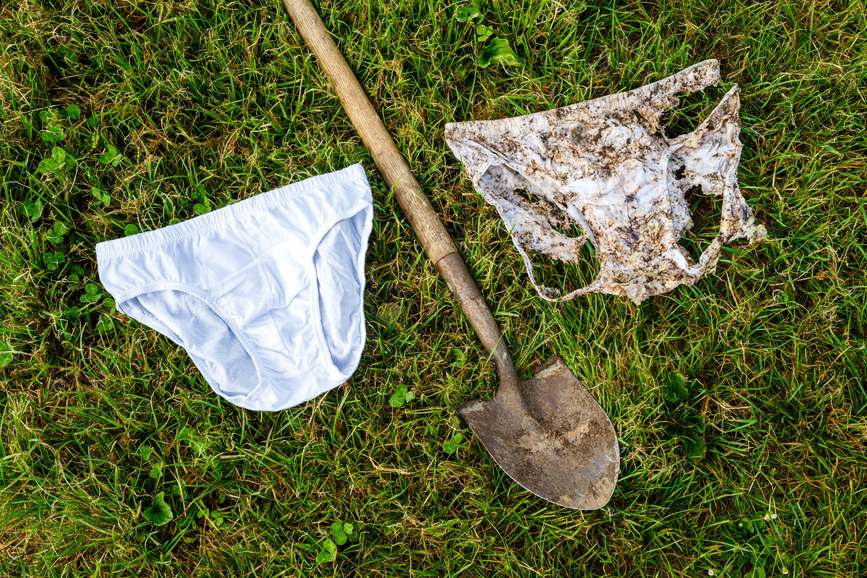 A new pair of white cotton briefs next to a pair torn apart after two months in soil, with a spade on the grass