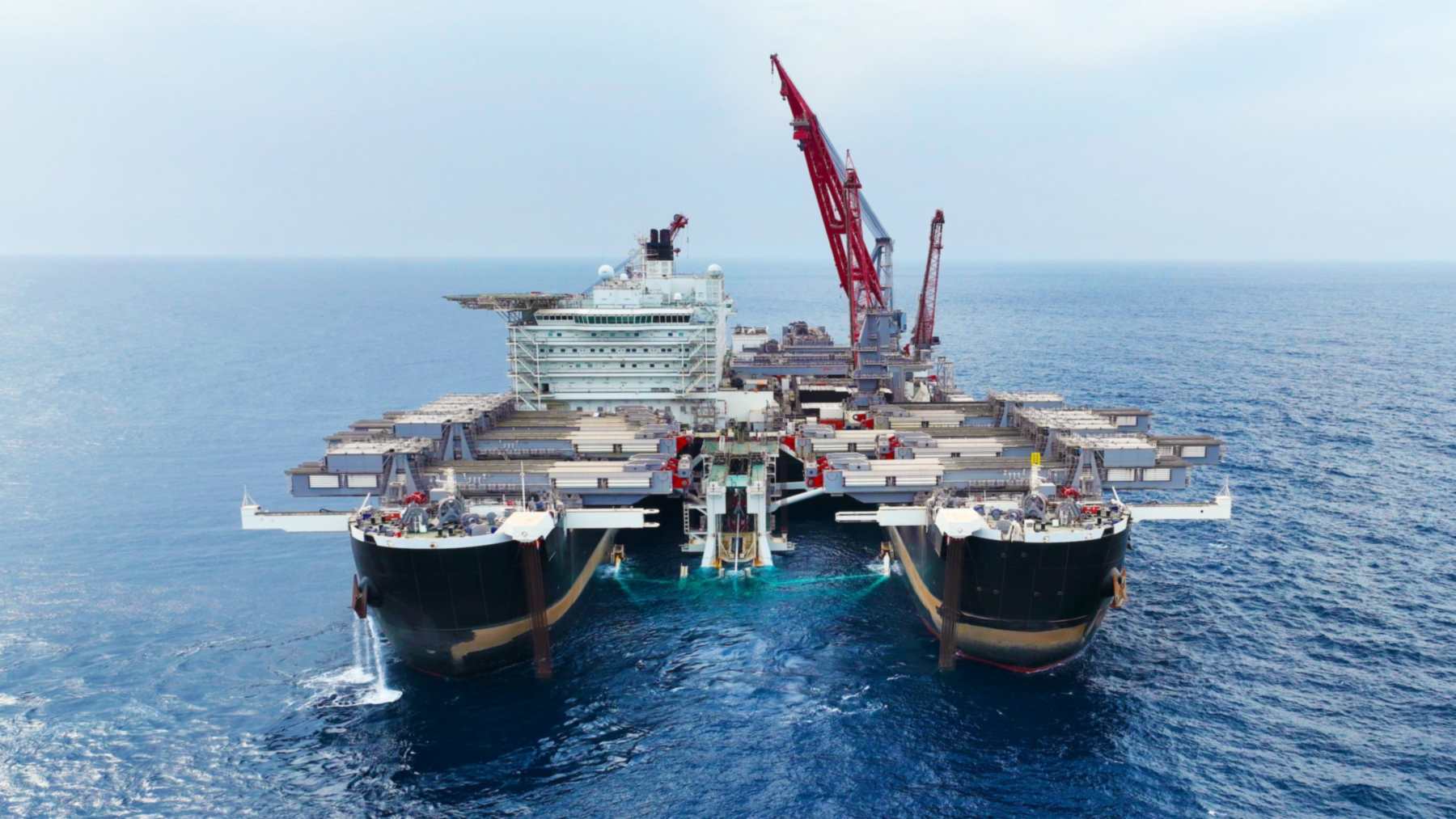Bow view of Allseas' Pioneering Spirit at sea, showing its two black hulls, the slot between them and a red crane rising behind the accommodation block