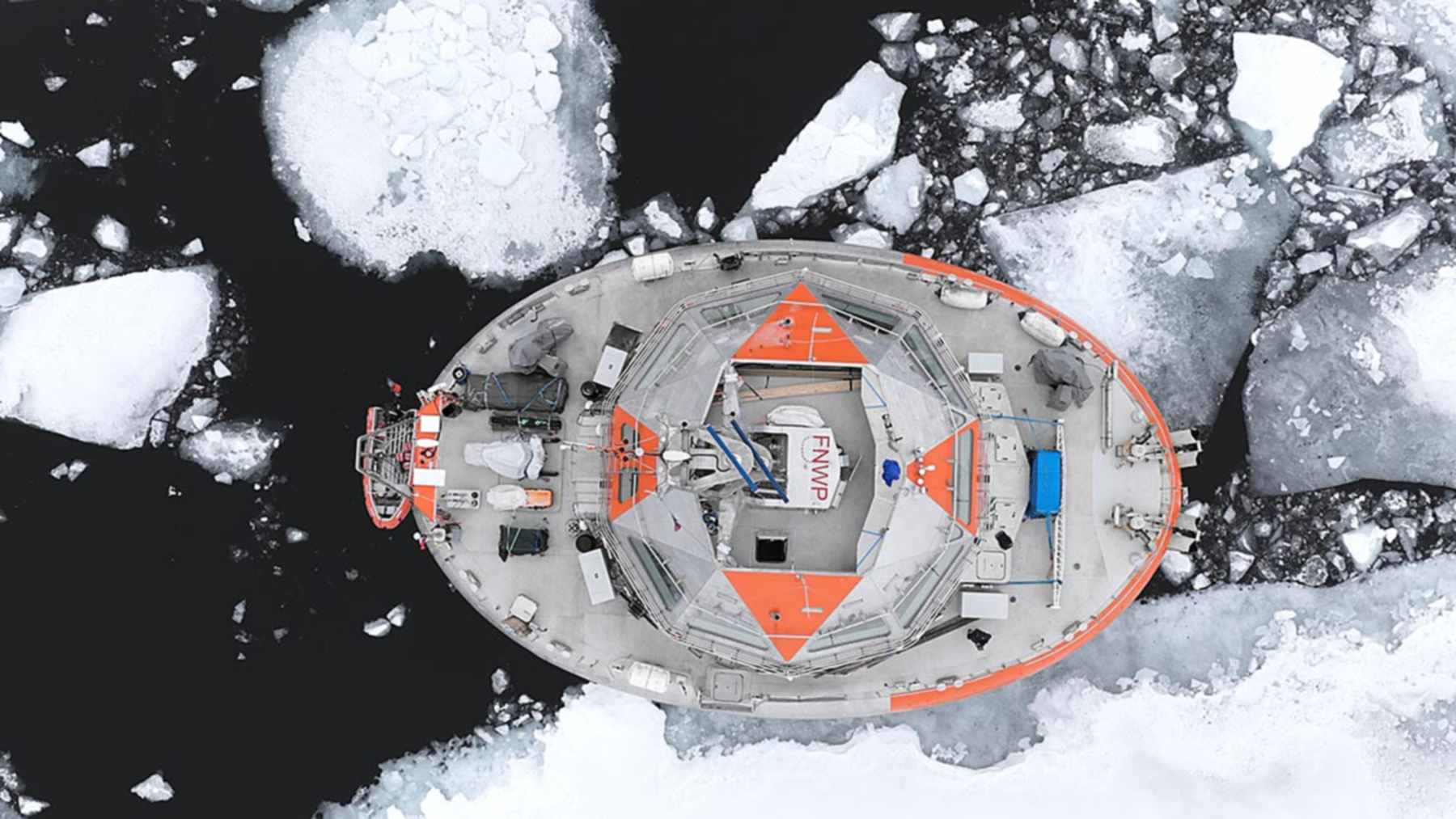 Aerial view of Tara Polar Station, a rounded orange and gray research vessel, surrounded by broken sea ice in the Arctic Ocean
