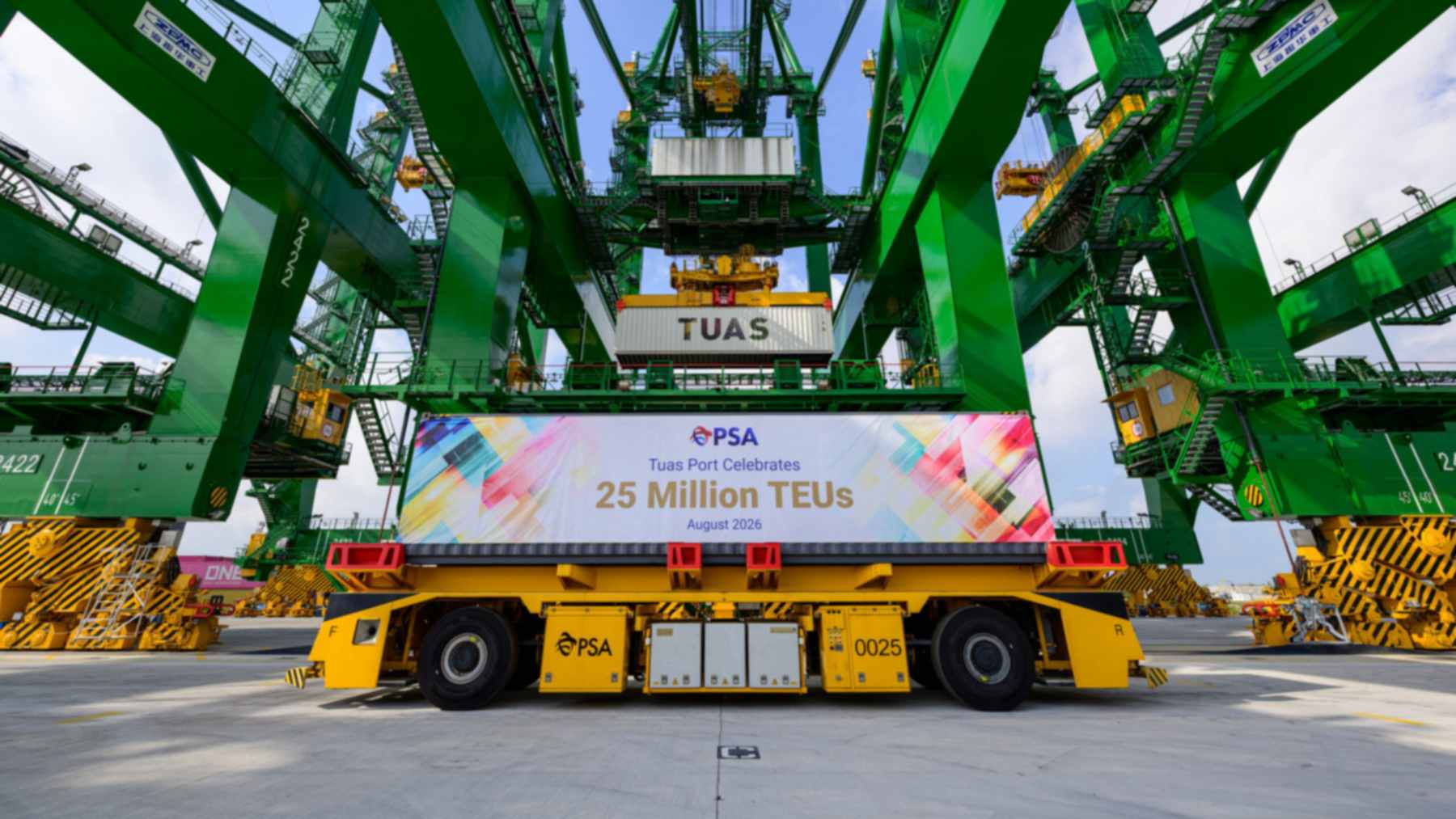 A yellow driverless container vehicle with a banner marking 25 million TEUs parked under green quay cranes at Tuas Port
