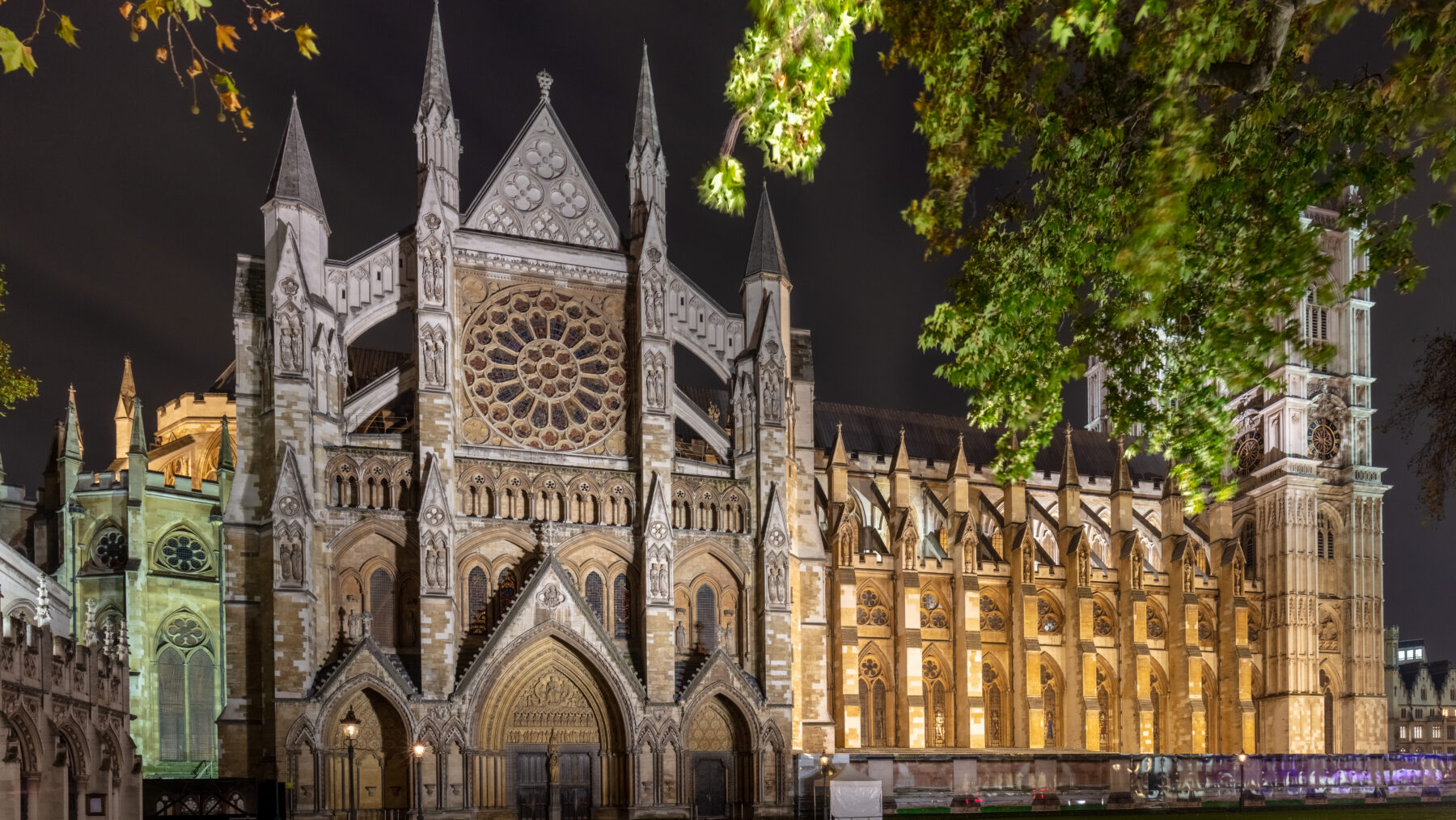 Westminster Abbey's north front lit up at night, with its large rose window and Gothic gables framed by autumn leaves.