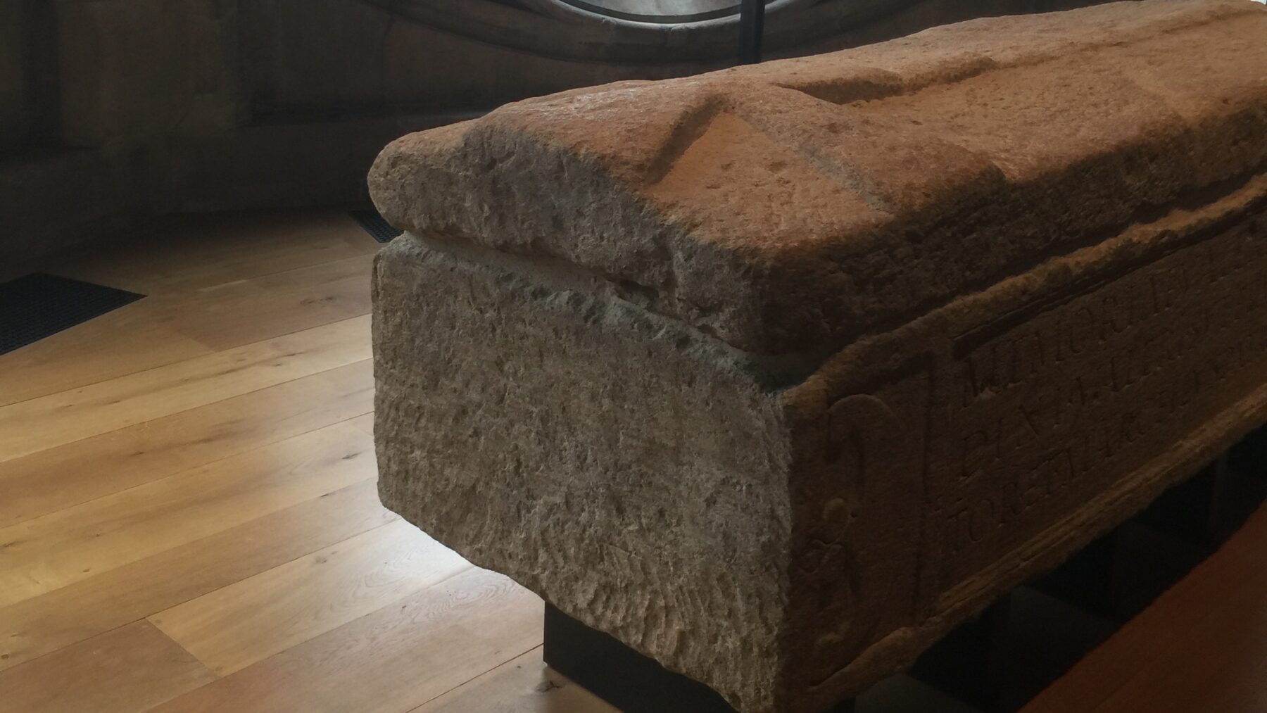 Weathered limestone Roman sarcophagus with a ridged lid and carved side panel, displayed in front of a Gothic rose window at Westminster Abbey.